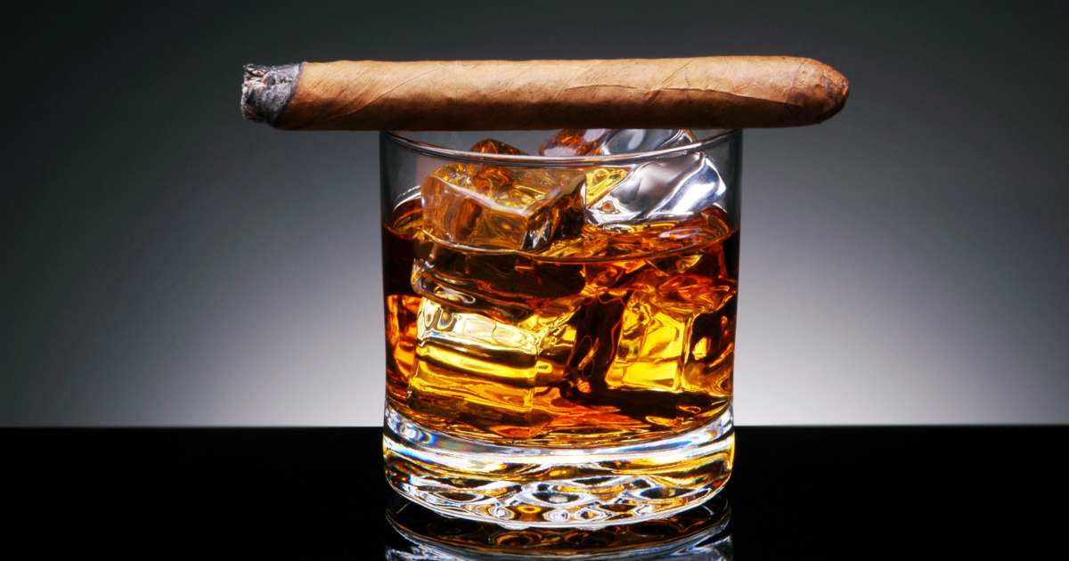 A cigar and a glass of bourbon sit on a wooden table, with a warm amber light illuminating the scene. The cigar has a dark wrapper and is partially lit, with smoke rising from the tip. The glass of bourbon is half-full and has a rich, caramel color. The table has a black surface.
