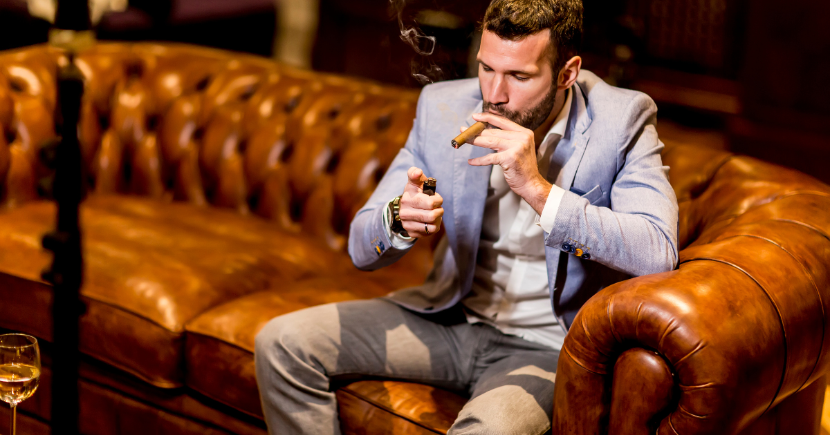 A man is sitting on a brown leather couch, holding a cigar in one hand and a lighter in the other. He wears a light suit, a light-colored shirt, and no tie. The cigar has a dark, oily wrapper and is unlit, with a cleanly cut foot. The man appears focused as he applies the flame from the lighter to the tip of the cigar, with a small flame visible. The background is out of focus, with the brown leather couch and some decorative elements visible.