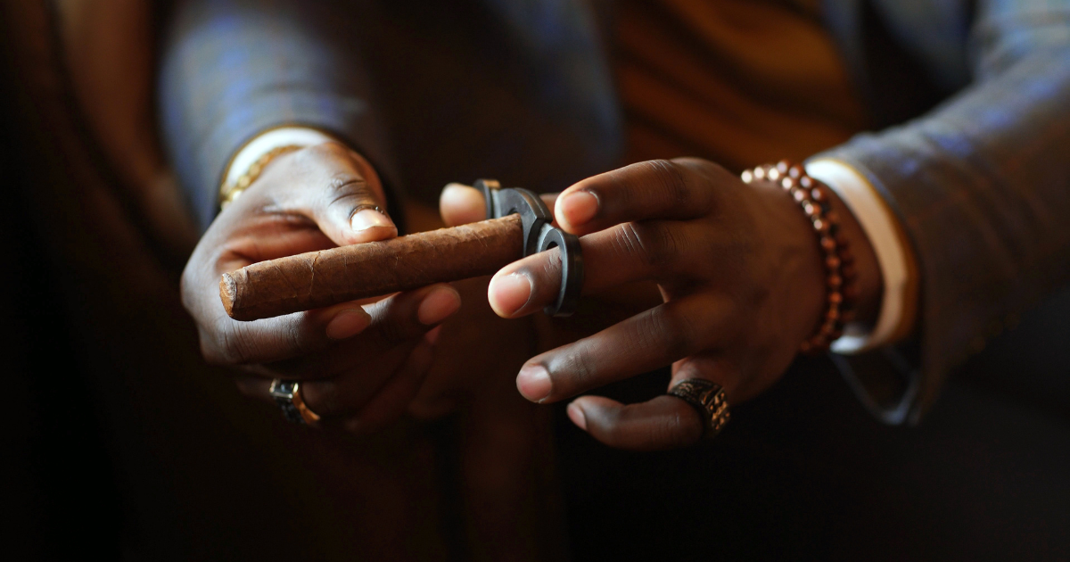 A man's hand holding a handheld cigar cutter, with a cigar positioned in the opening. The blades of the cutter are visible, ready to make a cut. The man's fingers are positioned to squeeze the cutter and make the cut. The background features a wooden surface.