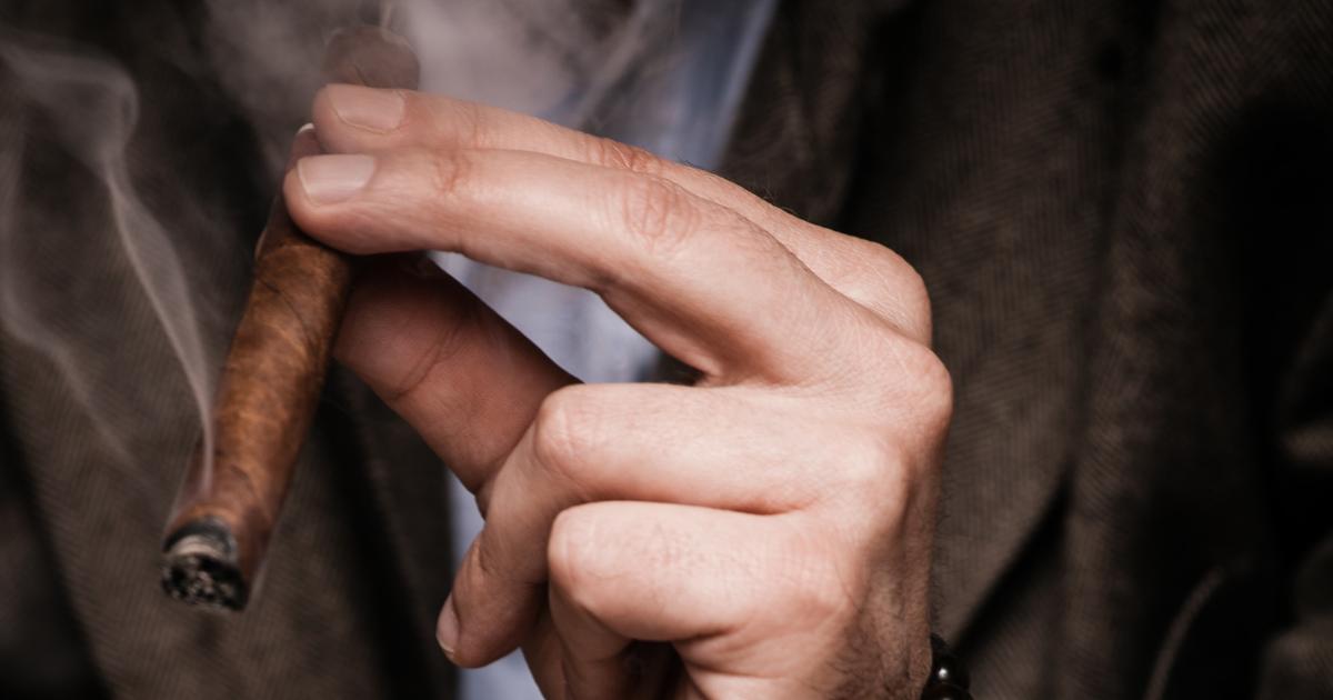 A man in a suit holding a cigar in his right hand, with smoke wafting up from the end. He has a serious expression and is looking off to the side.