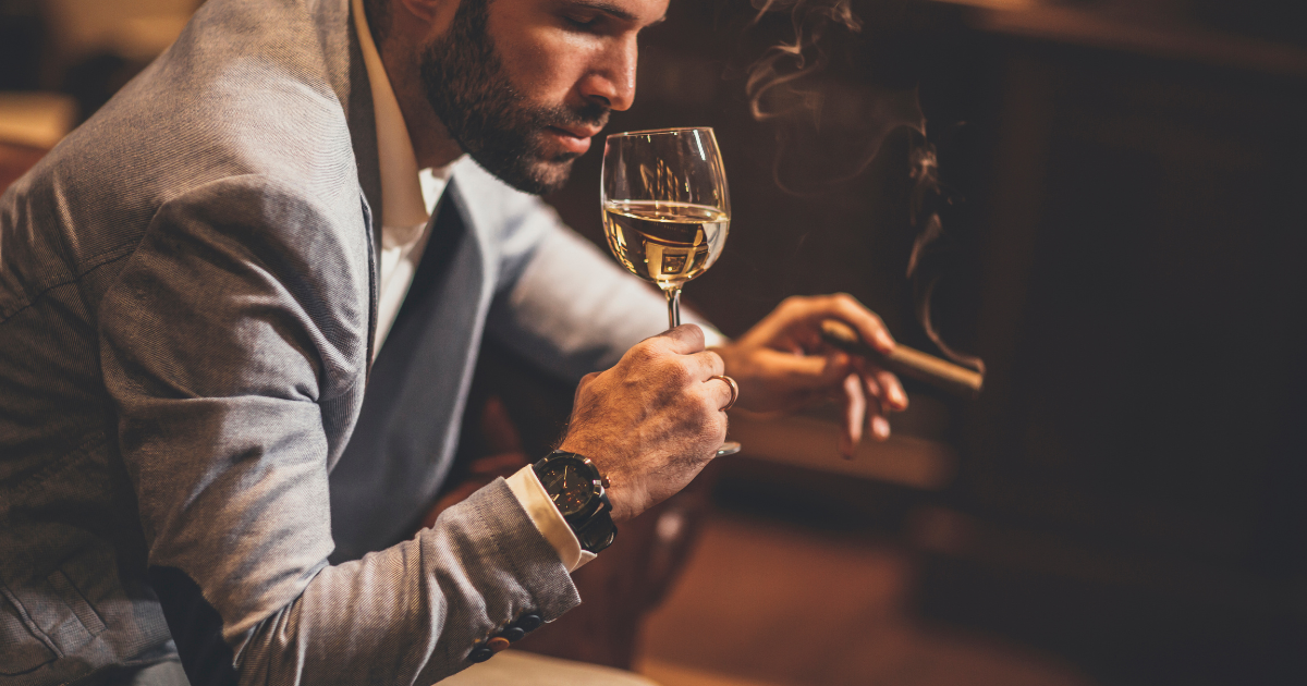 A man sits in a leather armchair holding a glass of white wine in one hand and a cigar in the other. He wears a suit and tie and appears to be enjoying the moment.